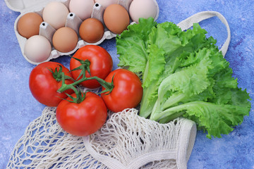 Fresh salad in a light mesh bag, red tomatoes and chicken eggs in a cardboard box on a blue background. View from above. Healthy food, eco, diet, organic food concept.