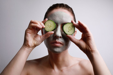 Portrait of young woman with a face mask on holding a cucumber slices in front of one eye. Beauty...