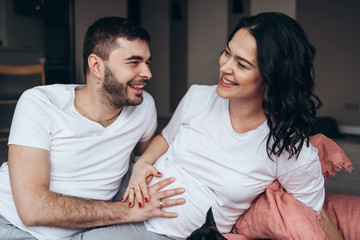 Young pregnant long hair woman with her husband at home. Man with beard is touching his wife belly and smile to her. Small black dog on a bed Future parents feeling their future baby through the belly