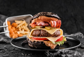 Black burger with beaf steak, lettuce, tomatoes, slice of cheese, ham, pastrami and sauce on slate black background, close up