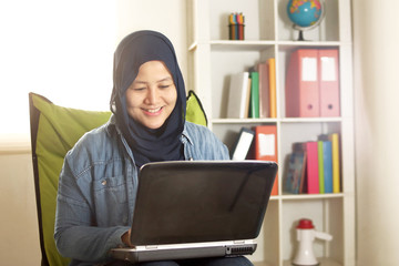 Portrait of happy smiling Asian muslim woman working with laptop in her bedroom, casual modern lifestyle