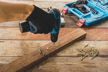 The carpenter works with an electric screwdriver, drilling the wood with a screw on the desk.
