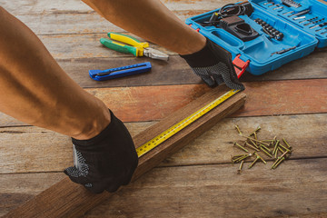 A carpenter is using a tape measure to measure the length of wood on the worker's desk floor To...