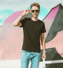 Young handsome guy is standing on a graffiti wall background. Man is wearing black, empty basic t-shirt without logo. Mock-up with light flare.