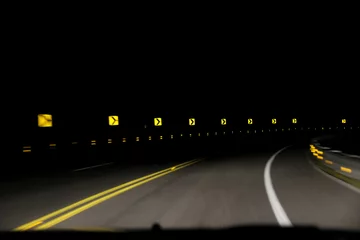 Foto op Plexiglas A series of yellow reflective 'sharp turn' signs being lit up by the lights of an oncoming car. The pitch black night sky is in contrast with the signs and street. Concept of speed and car accidents. © Thomas