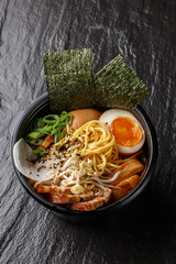 Asian noodle soup, ramen with pork, tofu, kimchi, vegetables and egg in black bowl. Slate...