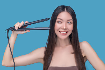 Young asian woman holding hair straightening iron and looking happy