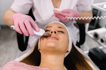 Obraz na płótnie Canvas Cosmetologist,beautician in white gloves making facial treatment with ultrasonic spatula to young woman,face skin scrubber treatment with ultrasonic spatula,facial cleansing procedure in beauty salon