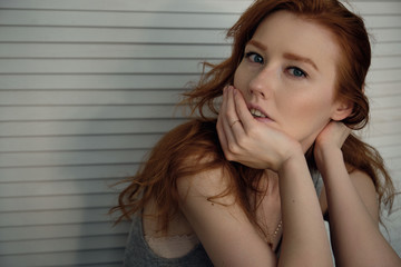 Red-haired girl with freckles sits against a white wooden screen and looks into the frame, propping her chin with her hand