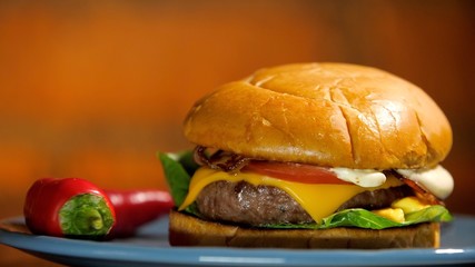 Juicy tasty burger lies on a plate with red chili peppers. Unhealthy quarantined foods cause obesity in America. Bun, fried cutlet, green juicy lettuce, crispy bacon, slice of cheese and tomato.
