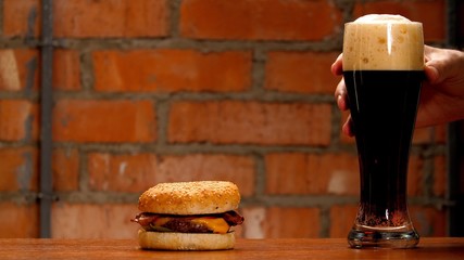 Man's hand holds a glass of cold black beer. Tasty beef burger with cheese and golden bun on wooden table on brick wall background. Unhealthy quarantine foods. 