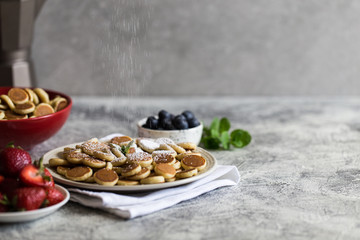 Tiny pancakes for breakfast. Cereal pancakes with blueberries, bananas, strawberries on grey background. Trendy food. Copy space for text or design
