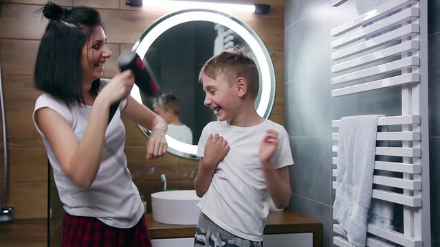 Cheerful lucky playful mother and child having fun in the bathroom while drying hair with hair dryer