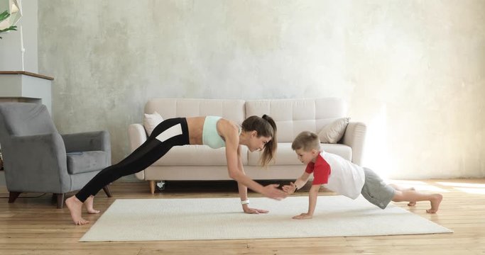 Mother and son are training together in plank exercise at home gives five each other. Sporty fit family doing fitness aerobic exercises in living room. Home workout, training and wellness concept.