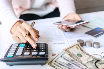 Young Asian woman holding smart phone while using calculator for business financial accounting calculate money bank loan rent payments manage expenses finances taxes doing paperwork concept, close up.