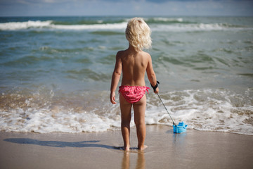 Happy little girl plays with a toy boat in the summer on the seashore. Family holidays with children by the sea. A child plays by the water. Blue toy boat.