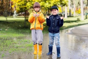 Two cheerfull friendly boys in rubber boots standing in puddle in park showing thumbs up gesture. Concept of social adaptation of autistic children. Friendship with peers. Full length portrait.
