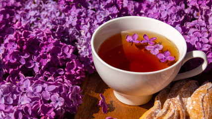 white porcelain cup with tea and a crispy bun among flowering branches of lilac...