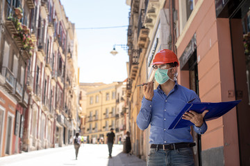 Ingegnere con casco arancione e camicia blu indossa una mascherina protettiva mentre parla al...