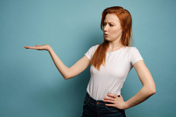 The red-haired girl stands on a blue background in a white T-shirt, points with the palm to the side and frowns looks there