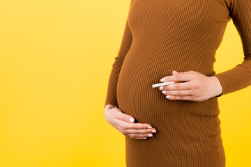 Close up of pregnant woman in brown dress with a cigarette against her belly at yellow background....