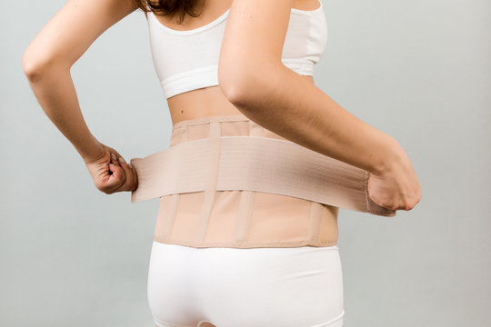 Back view of pregnant woman in underwear putting on supporting bandage to reduce backache at gray background with copy space. Cropped image of orthopedic abdominal support belt concept