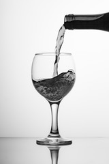 Black-white image of a bottle of wine and a glass. Pouring wine into a glass goblet on a high leg. Flowing liquid, wine drink, jet with waves and bubbles falling to the glass.