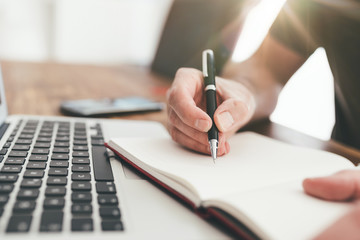 close-up of person taking notes with pen on paper in front of laptop computer