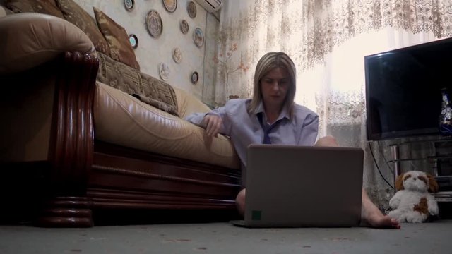 A young woman sitting on the floor of her house works behind a laptop.