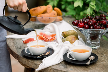 Tea ceremony, pouring tea from a teapot into a tea cup on a background of beautiful nature, fresh cherry and croissant. Hot cup of tea on rustic table. 