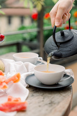 Tea ceremony, pouring tea from a teapot into a tea cup on a background of beautiful nature, fresh cherry and croissant. Hot cup of tea on rustic table. 