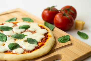 Homemade classic Italian Napoli pizza with tomato sauce, mozzarella cheese and basil leaves: pizza Napoletana. It is on a wooden cutting board surrounded by tomatoes, cheese and basil leaves