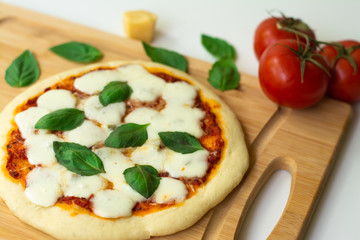 Homemade classic Italian Napoli pizza with tomato sauce, mozzarella cheese and basil leaves: pizza Napoletana. It is on a wooden cutting board surrounded by tomatoes, cheese and basil leaves