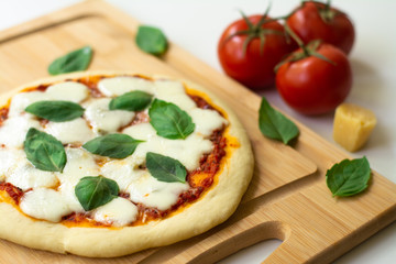 Homemade classic Italian Napoli pizza with tomato sauce, mozzarella cheese and basil leaves: pizza Napoletana. It is on a wooden cutting board surrounded by tomatoes, cheese and basil leaves