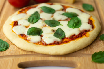Homemade classic Italian Napoli pizza with tomato sauce, mozzarella cheese and basil leaves: pizza Napoletana. It is on a wooden cutting board surrounded by tomatoes, cheese and basil leaves