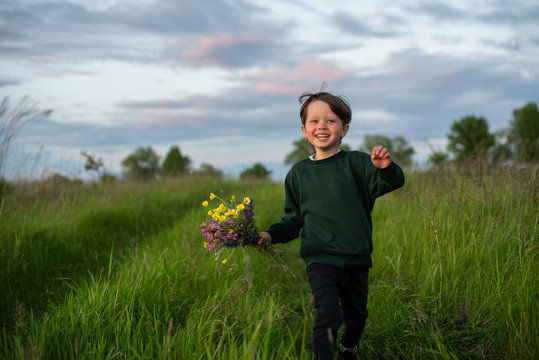 Happy little boy runs with a bouquet of wildflowers among tall green grass..