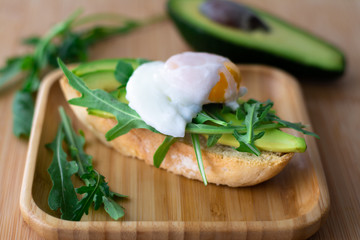 Stylish healthy breakfast of avocado, arugula and poached egg toast on a wooden surface with half an avocado and arugula in the background