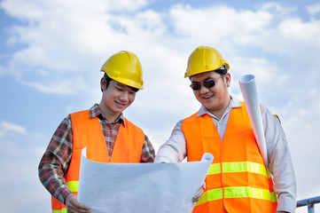 Two Asian engineers or technicians stand to hold a blueprint with a smile.