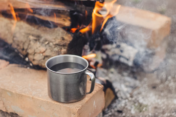 Metal cup of coffee water standing on the orange clay brick near the flame bonfire with firewood and smoke in spring. Outdoor activity bbq.