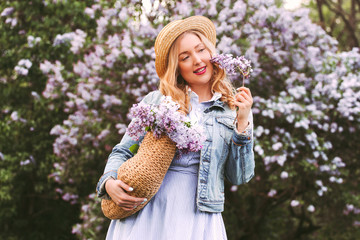 Portrait cheerful beautiful young hipster woman in denim dress, jacket smell lilac flower, smile in countryside park. Happy carefree blonde girl relax, have fun on blossom lilac garden. Spring allergy