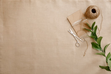 Top view image fashion designer background concept.Flat lay sewing scrapbooking accessory.Scissors, skein of twine, vintage envelope on brown linen fabric at home studio. Space for design mockup - obrazy, fototapety, plakaty