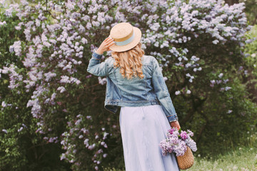 Back view stylish young beautiful blonde woman in denim jacket, dress, straw hat posing on blossom lilac garden. Happy attractive hipster girl walk, have fun in countryside park. Enjoy summer nature