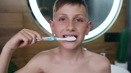 Front view of handsome smiling light haired boy which looking at camera while brushing his teeth in the bathroom