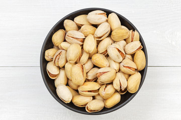 Fried pistachio nuts with salt in a black bowl on a white wooden table. Top view.