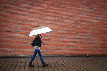Trendy woman with umbrella is walking next to red brick wall in rain