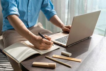 Asian businessman in blue shirt take note while looking at laptop on wood table with morning sunlight. Concept for working from home, e-learning during stay home and social distance against Covid 19 