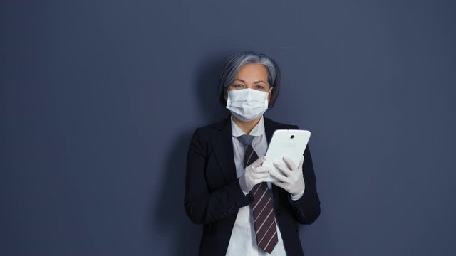 Grey haired businesswoman in protective mask and gloves holding and looking at tablet standing in the dark blue wall background . Health care concept. Business concept. Prores 422