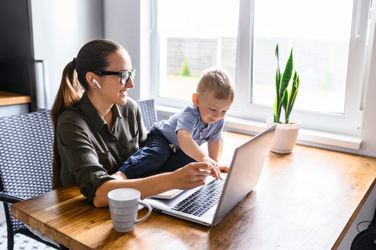 Stay home. Mother works from home, she sits in the kitchen and using laptop for remote work, her cute baby boy sits on the table