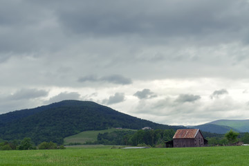 A rural scene depicting an old barn in a hay pasture with storm clouds overhead and mountains in the background.