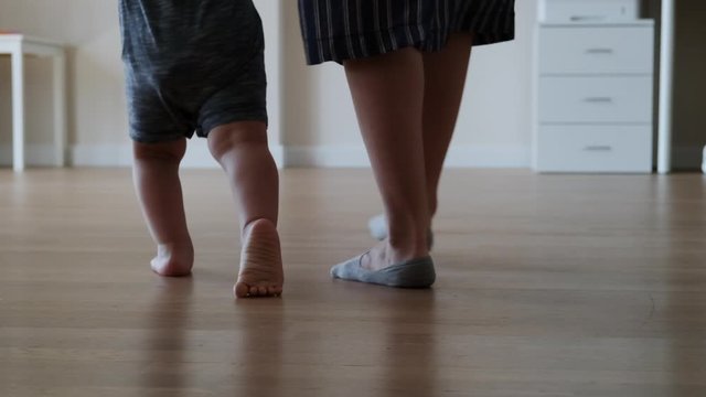 Little kid feet with mother walking on floor, close-up. Baby learning to walk at home. Baby first steps. Slow motion